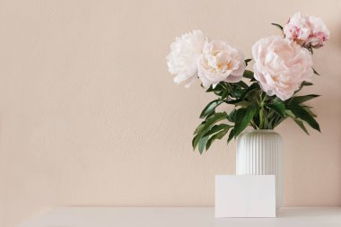 Bahar, yaz çiçekleri natürmort. Beyaz ahşap masadaki porselen vazoda pembe şakayık çiçekleri. Boş tebrik kartı maketi sahnesi. Arka tarafta çıplak duvar var. Boş kopya psace, insan yok, ön görünüm.