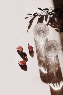 Yaz hala hayat sahnesi. İki bardak su, uzun gölgeli kokteyller, incir meyveleri ve bej masa arkasında güneş ışığı altında zeytin yaprakları. Dikey düzlük, üst görünüm. Doğum günü kavramı.