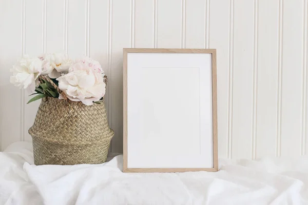 Beige blank wooden picture frame mockup. Artistic table still life composition with pink peony flowers in straw basket. White wooden wall background. Empty copyspace, rustic design. — Stock Photo, Image