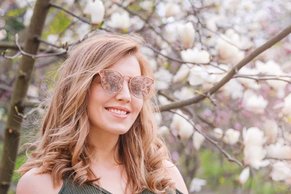 Ung blond kvinna nära blommande magnolia blommor träd i vår park på solig dag. Magnoliaträd. Vacker glad flicka njuter av lukten i en blommande vårträdgård — Stockfoto