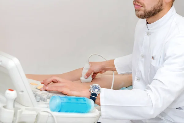 Beyaz üniformalı bir doktor, teşhis cihazıyla kadın bir hastanın bacaklarındaki cihazı kullanarak ultrason yapar. Cerrah, filebolog. Moder Kliniği 'nde çalışıyor. Hastanede tıbbi aletler.