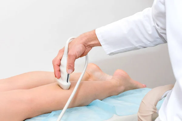 Doctor in white uniform with diagnostic equipment performs an ultrasound using a device on the legs of a female patient. Surgeon man, phlebologist working in moder clinic. Medical tools in hospital. — Stock Photo, Image