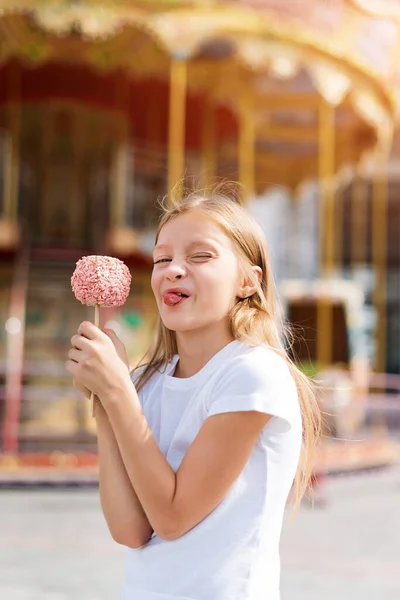 Cute little girl eating candy apple and posing at fair in amusement park