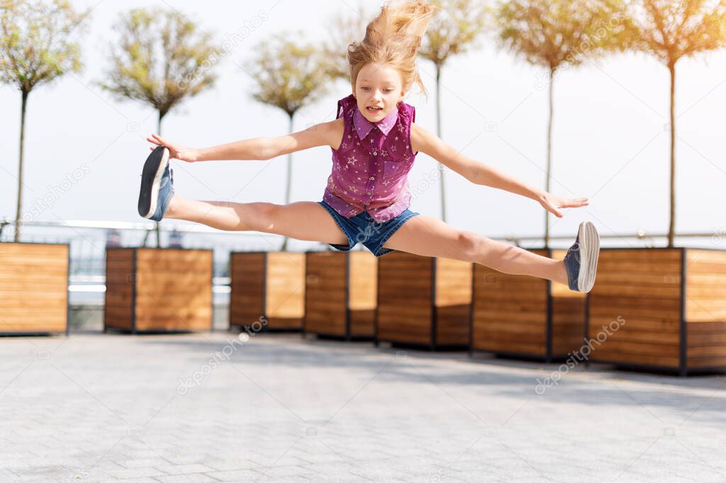 Active kid girl gymnast jumping on the street, doing splits. Stretching exercise, straight angle pose in the air. Young girl acrobat. The girl is engaged in gymnastics