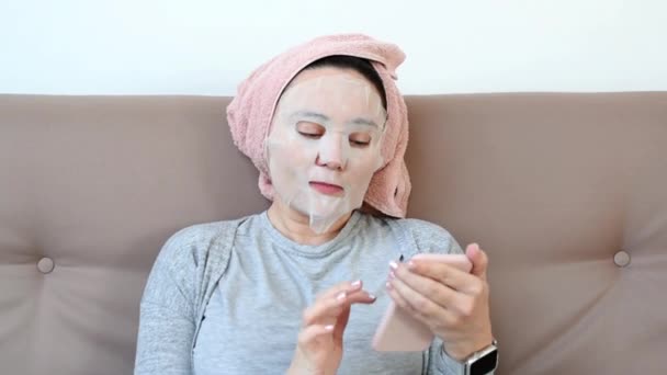 Giovane 30s donna con maschera cosmetica facciale sul suo viso utilizzando il telefono cellulare a casa. Donna beauty routine, riposo a casa. Isolamento autonomo, quarantena domestica, allontanamento sociale — Video Stock