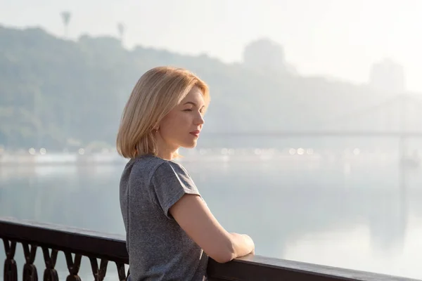 Young blonde woman enjoying city view from the bridge. Lovely woman spending time on the river embankment at sunset. Soft backlit. Lifestyle relax recreation weekend concept.