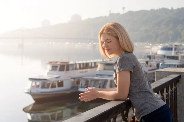Young blonde woman enjoying city view from the bridge. Lovely woman spending time on the river embankment at sunset. Soft backlit. Lifestyle relax recreation weekend concept.