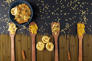 Tahta spatulalı çeşitli makarnalar ve dokulu siyah zemin üzerinde mavi bir seramik kase, üst görünüm, metin alanı, düz makarna