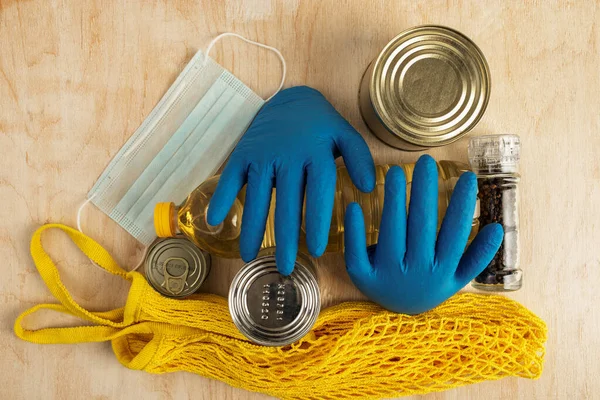 medical protection against covid-19 virus and long-term storage products on a light wooden background. medical mask, volumetric medical gloves and string bag on a bottle of sunflower oil and canned food, top view, flat lay food