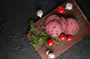 Siyah bir tablo ve bir kahverengi tahta üzerinde füme salam. Biberiye, sarımsak ve biber ve domates kiraz, dereotu, maydanoz ile sosis.