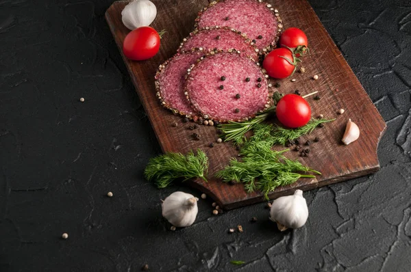 Gerookte salami op een zwarte lijst en een bruin bord. Worst met knoflook en peper en tomaat cherry, dille, rozemarijn, peterselie. — Stockfoto