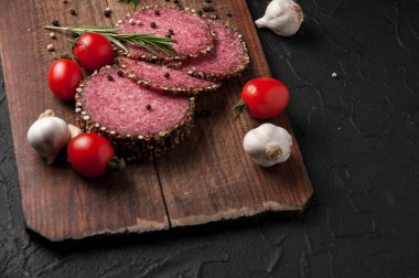 Siyah bir tablo ve bir kahverengi tahta üzerinde füme salam. Biberiye, sarımsak ve biber ve domates kiraz, dereotu, maydanoz ile sosis.