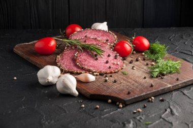 Siyah bir tablo ve bir kahverengi tahta üzerinde füme salam. Biberiye, sarımsak ve biber ve domates kiraz, dereotu, maydanoz ile sosis.