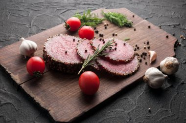 Siyah bir tablo ve bir kahverengi tahta üzerinde füme salam. Biberiye, sarımsak ve biber ve domates kiraz, dereotu, maydanoz ile sosis.