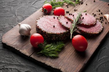 Siyah bir tablo ve bir kahverengi tahta üzerinde füme salam. Biberiye, sarımsak ve biber ve domates kiraz, dereotu, maydanoz ile sosis.