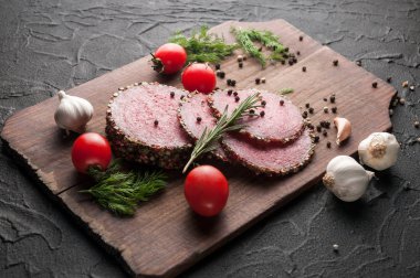 Siyah bir tablo ve bir kahverengi tahta üzerinde füme salam. Biberiye, sarımsak ve biber ve domates kiraz, dereotu, maydanoz ile sosis.