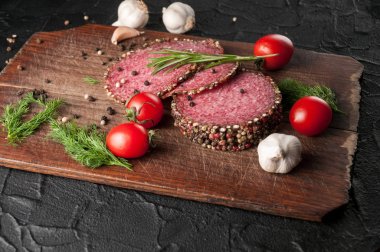 Siyah bir tablo ve bir kahverengi tahta üzerinde füme salam. Biberiye, sarımsak ve biber ve domates kiraz, dereotu, maydanoz ile sosis.