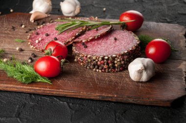 Siyah bir tablo ve bir kahverengi tahta üzerinde füme salam. Biberiye, sarımsak ve biber ve domates kiraz, dereotu, maydanoz ile sosis.