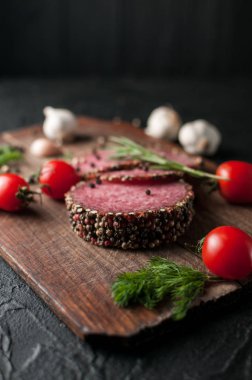 Siyah bir tablo ve bir kahverengi tahta üzerinde füme salam. Biberiye, sarımsak ve biber ve domates kiraz, dereotu, maydanoz ile sosis.