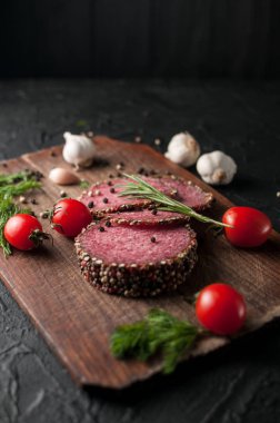 Siyah bir tablo ve bir kahverengi tahta üzerinde füme salam. Biberiye, sarımsak ve biber ve domates kiraz, dereotu, maydanoz ile sosis.