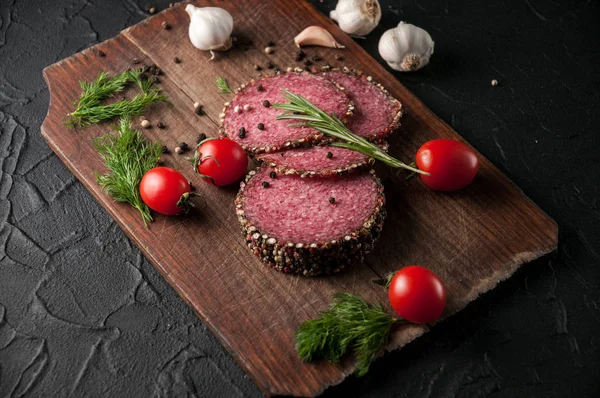 Siyah bir tablo ve bir kahverengi tahta üzerinde füme salam. Biberiye, sarımsak ve biber ve domates kiraz, dereotu, maydanoz ile sosis.