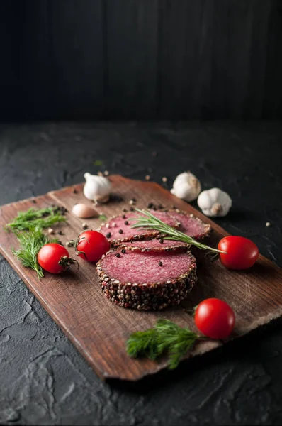 Siyah bir tablo ve bir kahverengi tahta üzerinde füme salam. Biberiye, sarımsak ve biber ve domates kiraz, dereotu, maydanoz ile sosis.