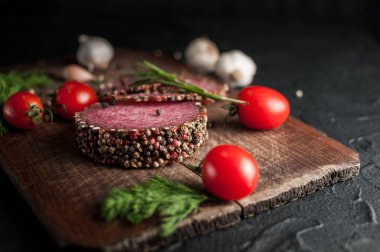 Siyah bir tablo ve bir kahverengi tahta üzerinde füme salam. Biberiye, sarımsak ve biber ve domates kiraz, dereotu, maydanoz ile sosis.