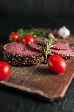 Siyah bir tablo ve bir kahverengi tahta üzerinde füme salam. Biberiye, sarımsak ve biber ve domates kiraz, dereotu, maydanoz ile sosis.