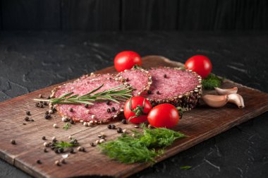 Siyah bir tablo ve bir kahverengi tahta üzerinde füme salam. Biberiye, sarımsak ve biber ve domates kiraz, dereotu, maydanoz ile sosis.