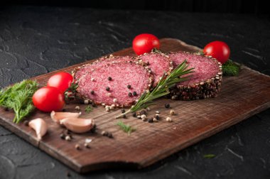 Siyah bir tablo ve bir kahverengi tahta üzerinde füme salam. Biberiye, sarımsak ve biber ve domates kiraz, dereotu, maydanoz ile sosis.