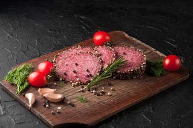 Siyah bir tablo ve bir kahverengi tahta üzerinde füme salam. Biberiye, sarımsak ve biber ve domates kiraz, dereotu, maydanoz ile sosis.