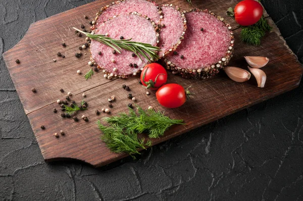 Gerookte salami op een zwarte lijst en een bruin bord. Worst met knoflook en peper en tomaat cherry, dille, rozemarijn, peterselie. — Stockfoto