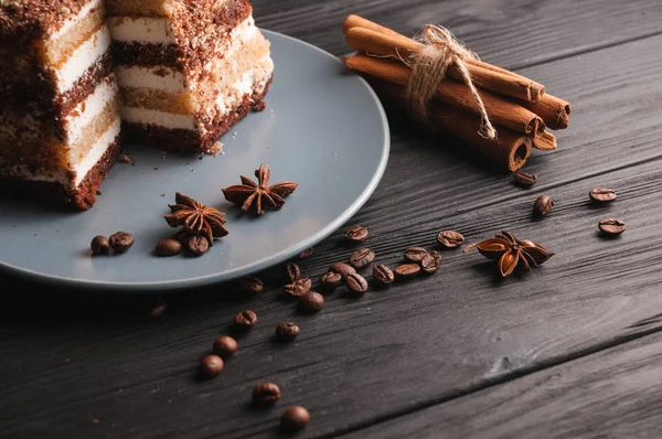 Tiramisú dulce de postre en un corte en un plato gris sobre un fondo de madera negro con especias: palitos de canela y badan, granos de café . — Foto de Stock