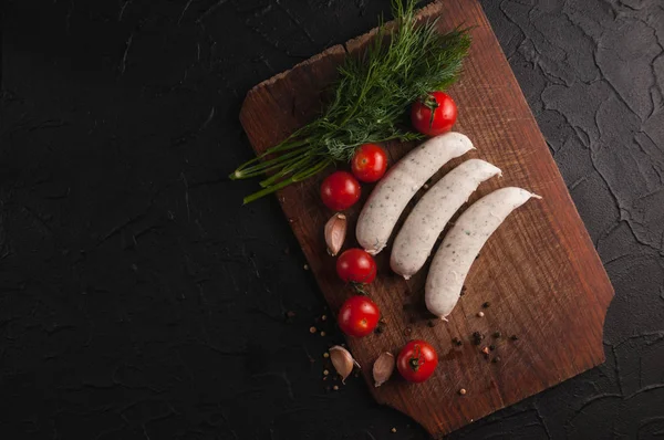 Drie rauwe worstjes op een houten bruin bord op een zwarte achtergrond met cherrytomaatjes, peterselie en dille, peper en knoflook. — Stockfoto