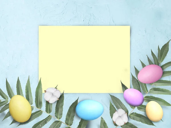 Mock up Easter flatlay with empty yellow blank, colorful eggs, cotton flowers and leaves on blue background. Greeting holiday composition with copy space.