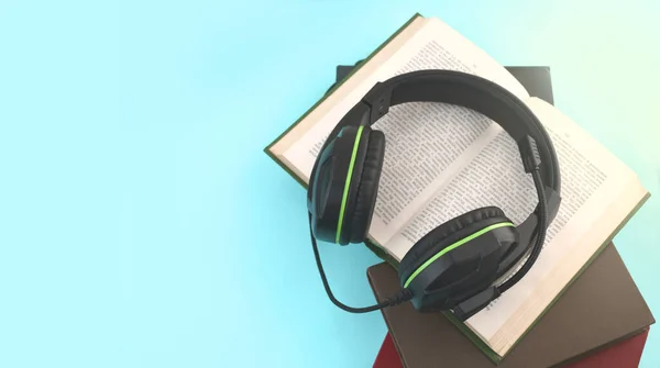 Audio book concept. Headphones, open book and pile of books on turquose background. Listening to book. Modern education. Top view, copyspace.