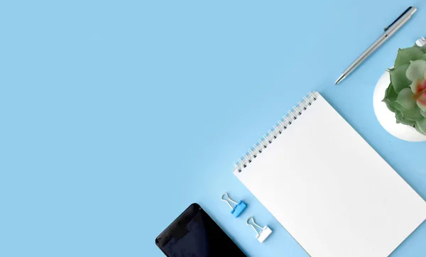 Minimalist office flatlay with empty notepad, smartphone, succulent and stationery on blue background.  Online education or workplace concept.  Banner / header with copy space