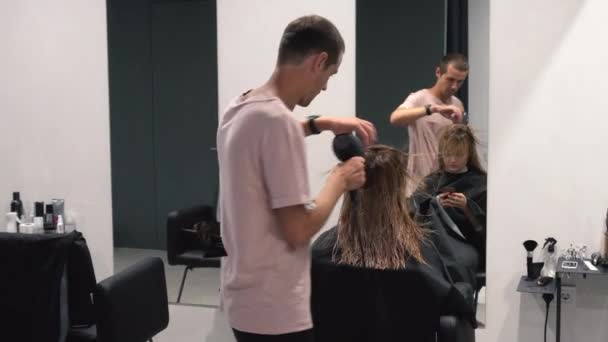 Belle femme jouant avec Smartphone dans le salon de coiffure tandis que le coiffeur professionnel coupe les cheveux avec ciseaux dans le salon de coiffure. Concept de beauté — Video