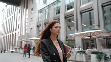 Kulaklıklı genç bir iş kadını iş merkezinin yakınında dizüstü bilgisayarla yürüyor.