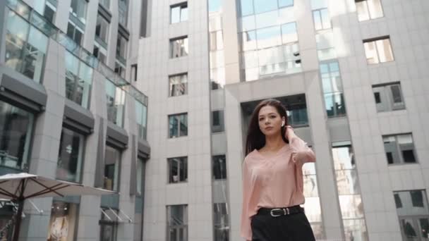 Молодая деловая женщина в наушниках, идущая рядом с бизнес-центром — стоковое видео