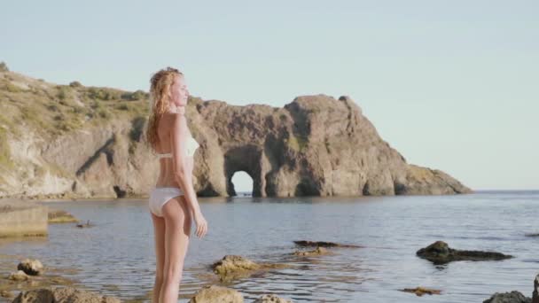 Fiatal felnőtt hölgy fehér fürdőruhában a tenger mellett áll — Stock videók
