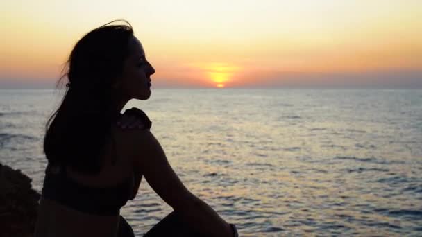Młoda dorosła dziewczyna siedzi w pobliżu morza, patrząc na zachód słońca — Wideo stockowe