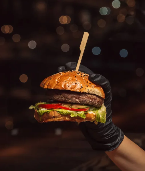 Mano en guantes de hamburguesa sosteniendo hamburguesa de carne en fondo negro — Foto de Stock