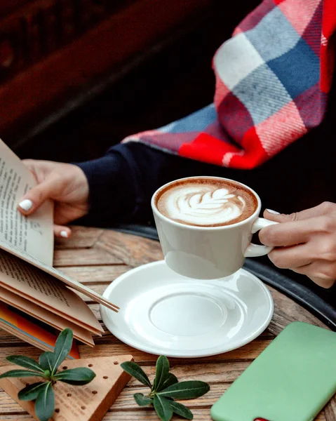 O femeie care ține o ceașcă de cappuccino și citește o carte — Fotografie, imagine de stoc