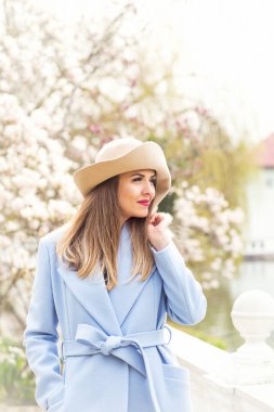 Mavi ceketli ve açık renk şapkalı genç, güzel bir kadın güneşli bir günde, çiçek açan bir Magnolia ağacının yanında, baharda parkta yürür.