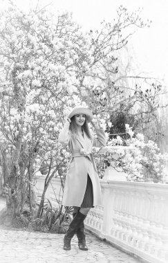 Güneşli bir günde, çiçek açan bir manolya ağacının yanında parka yürürken, şık bir ceket ve şapkayla genç, güzel bir kadının siyah ve beyaz fotoğrafı.