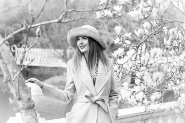 Güneşli bir günde, çiçek açan bir manolya ağacının yanında parka yürürken, şık bir ceket ve şapkayla genç, güzel bir kadının siyah ve beyaz fotoğrafı.