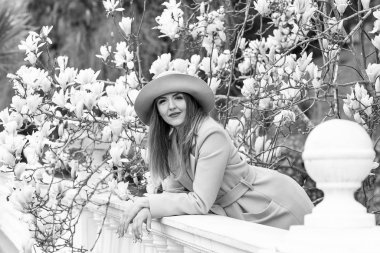 Güneşli bir günde, çiçek açan bir manolya ağacının yanında parka yürürken, şık bir ceket ve şapkayla genç, güzel bir kadının siyah ve beyaz fotoğrafı.