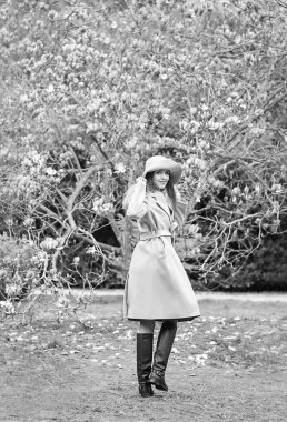 Güneşli bir günde, çiçek açan bir manolya ağacının yanında parka yürürken, şık bir ceket ve şapkayla genç, güzel bir kadının siyah ve beyaz fotoğrafı.