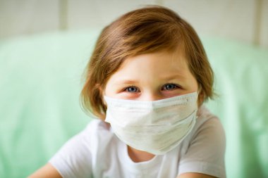 Küçük bir kız, tıbbi maske takıyor, evde oynuyor, karantinada, koronavirüs salgını sırasında, COVID-2019
